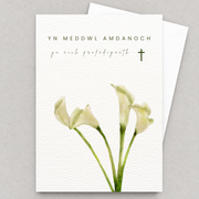 Cerdyn cydymdeimlad gyda lilïau gwyn a chroes fach, yn gorwedd yn fflat gydag amlen wen