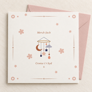 Welsh baby girl card with baby mobile and pastel flowers, lying flat with a blush pink envelope.