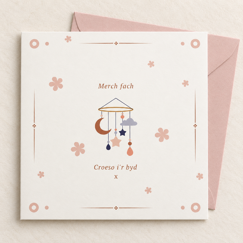 Welsh baby girl card with baby mobile and pastel flowers, lying flat with a blush pink envelope.