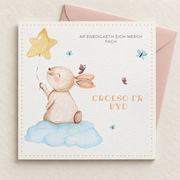 Welsh baby girl card with rabbit on a blue cloud holding a star balloon, lying flat with a blush pink envelope.