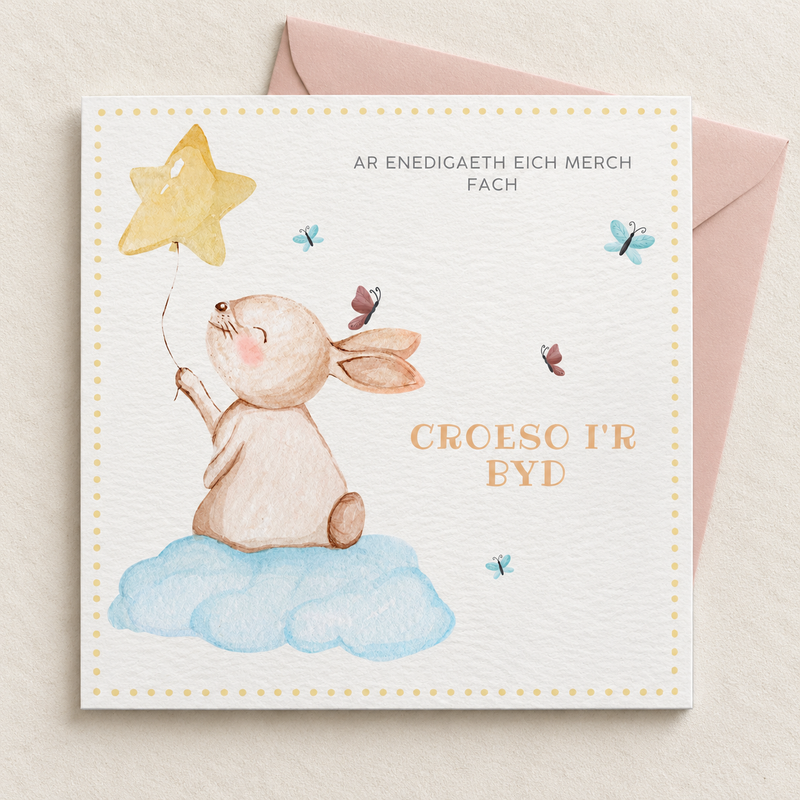 Welsh baby girl card with rabbit on a blue cloud holding a star balloon, lying flat with a blush pink envelope.