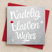 Cerdyn Nadolig Llawen Wyres gyda llythrennau gwyn a phlu eira ar gefndir llwyd, yn wastad gydag amlen goch / Welsh Christmas card lying flat with a red envelope